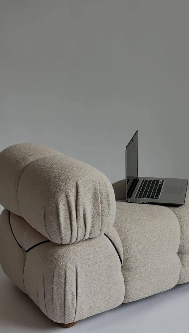 Sculptural boucle lounge chair with an open laptop resting on its arm against a soft grey wall